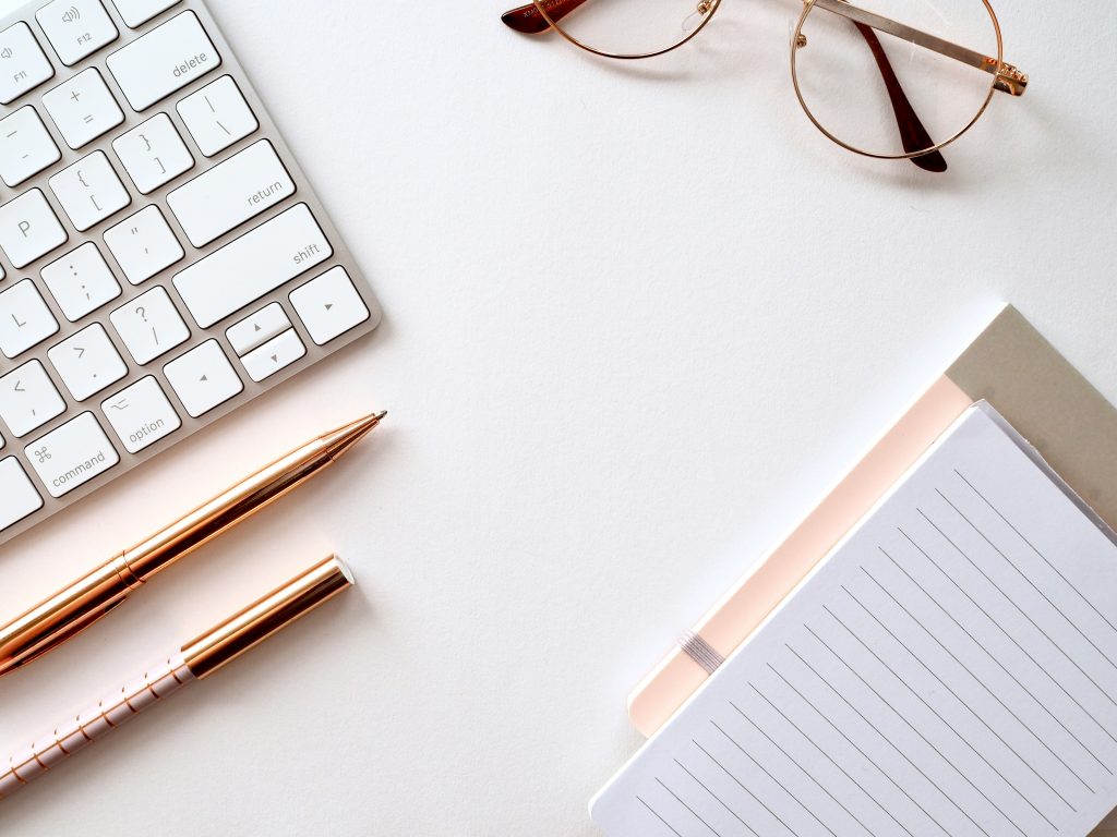 Rose gold aesthetic of a laptop keyboard, pens, a notebook, and glasses on a desk. Photo by Jess Bailey via Unsplash). 